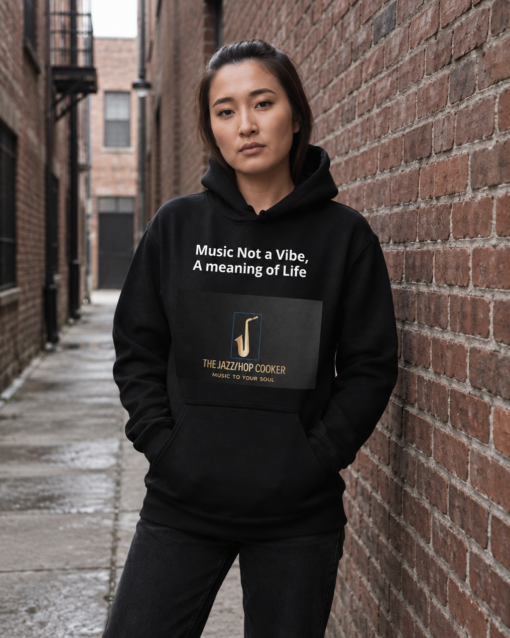A woman stands in a brick alley wearing a black hoodie with white text reading 'Music Not a Vibe, A meaning of Life' and a dark front graphic that says 'THE JAZZHIP COOKER' with a jazz note logo.