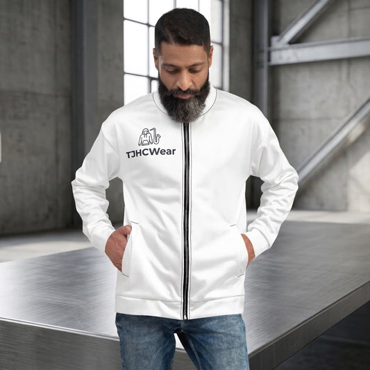 Man wearing a white TJHCWear unisex bomber jacket with a black full-length zipper, black chest jazz-and-saxophone logo, stand-up collar, long sleeves, and side pockets in an industrial studio setting.