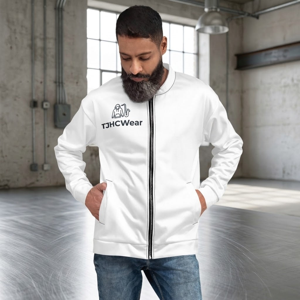 A man with a beard wearing a white zip-up jacket with a black front zipper and a black printed logo that reads 'TJHCWear' on the left chest, paired with blue jeans, standing in an industrial loft setting.